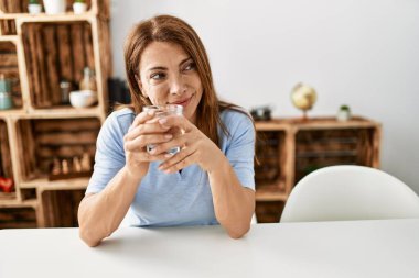Orta yaşlı beyaz bir kadın evdeki masada oturmuş su içiyor..