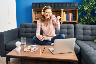 Young beautiful woman practicing painting looking at online tutorial screaming proud, celebrating victory and success very excited with raised arms 