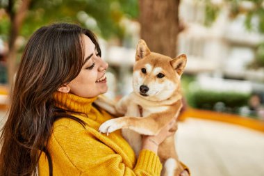 Güzel genç bir kadın parkta mutlu Shiba Inu köpeğine sarılıyor.