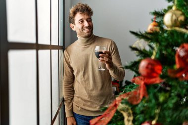 Genç İspanyol adam evde Noel ağacının yanında şarap içiyor.