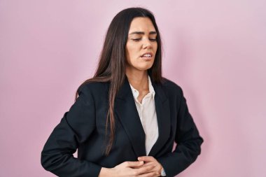 Genç esmer kadın pembe arka planda iş stili giyiniyor, elleri karnında, çünkü hazımsızlık, acı verici bir hastalık kendini iyi hissetmiyor. Ağrı kavramı. 