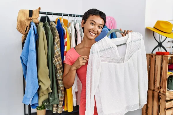Young hispanic woman customer smiling confident choosing clothes holding shirt at clothing store