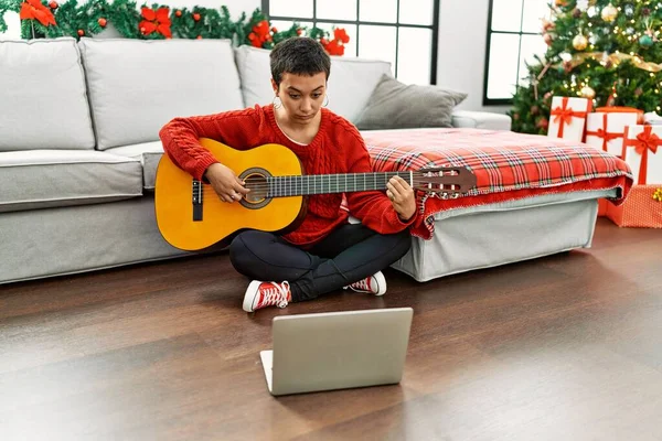 Young hispanic woman having online guitar class sitting by christmas tree at home