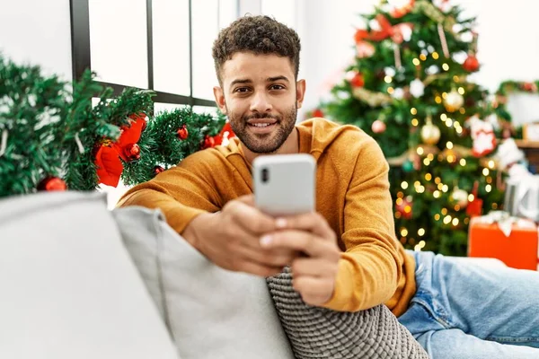 Νέοι Άραβας Άνθρωπος Χρησιμοποιώντας Smartphone Κάθεται Στον Καναπέ Από Χριστουγεννιάτικο — Φωτογραφία Αρχείου