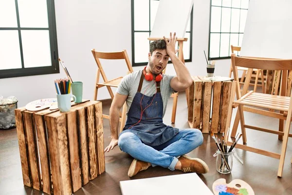 Young hispanic man sitting at art studio surprised with hand on head for mistake, remember error. forgot, bad memory concept.