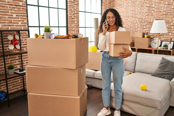 Genç Latin kadın akıllı telefondan konuşuyor. Yeni evinde paketleri tutuyor.