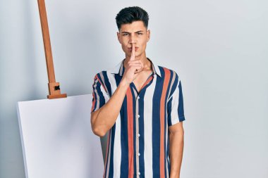 Genç İspanyol bir adam boş tuvalin yanında duruyor ve parmakları dudaklarında sessiz olmak istiyor. sessizlik ve gizli konsept. 