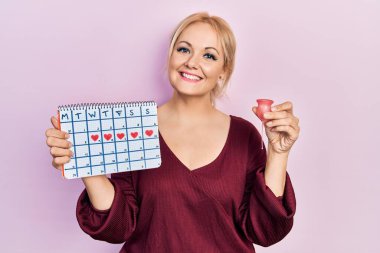 Kalp takvimi ve regl fincanı tutan genç sarışın kadın yüzünde mutlu ve havalı bir gülümsemeyle gülümsüyor. diş gösterme. 