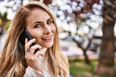 Genç sarışın kadın gülümsüyor. Parktaki akıllı telefondan konuşuyor.