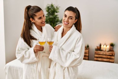 Bornoz giymiş iki kadın, güzellik merkezinde şampanyayla kadeh kaldırıyor.