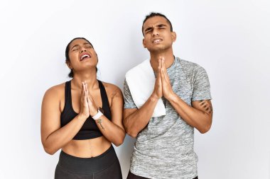 Spor kıyafetleri giyen genç Latin bir çift izole edilmiş arka planda duruyor. Elleri bağlı yalvarıyor ve dua ediyorlar. Yüzlerinde umut ifadesi var. Yalvarıyor. 