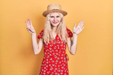 Beautiful caucasian woman with blond hair wearing summer hat showing and pointing up with fingers number eight while smiling confident and happy. 