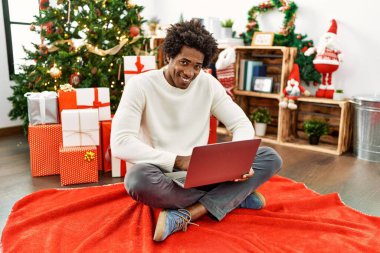 Genç Afro-Amerikan adam dizüstü bilgisayar kullanıyor evdeki noel ağacının yanında oturuyor..