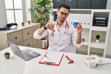 Sakallı, kulak pamuğu tutan, kızgın yüzlü, negatif işaretli, baş parmakları aşağıda, reddedilme konsepti olan genç İspanyol bir doktor. 