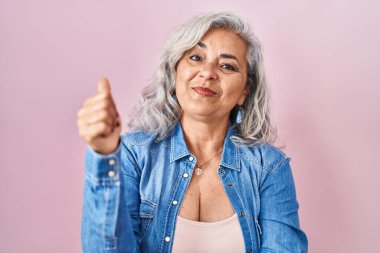 Pembe arka planda gri saçlı, gururlu, gülümseyen, baş parmak kaldıran orta yaşlı bir kadın. 