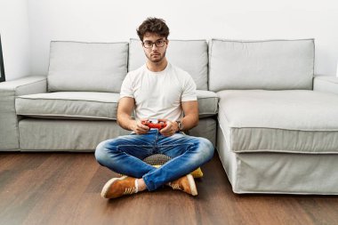 Hispanic man playing video game holding controller by the sofa thinking attitude and sober expression looking self confident 