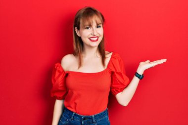 Kırmızı tişörtlü, neşeli gülümseyen, avuç içi kameraya bakan kızıl saçlı genç kadın.. 