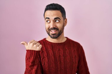 Sakallı, pembe arka planda günlük süveter giyen, gülümseyen, gülümseyen ve baş parmağıyla yüzünü gösteren genç bir İspanyol.. 