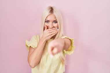 Pembe arka planda duran beyaz kadın sana gülüyor, eli ağzında kamerayı işaret ediyor, yüz ifadesi utanç verici. 