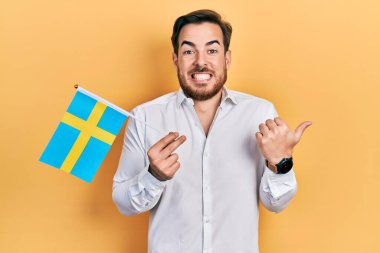 Sakallı yakışıklı, beyaz bir adam elinde İsveç bayrağı başparmağını kaldırmış, ağzı açık gülümsüyor. 