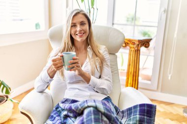 Genç sarışın kadın kahve içiyor. Evdeki koltukta oturuyor..