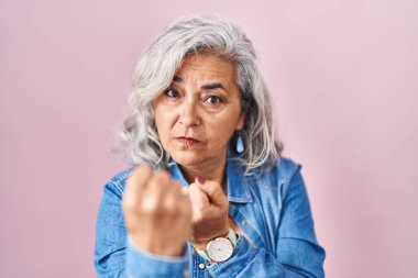 Pembe arka planda gri saçlı orta yaşlı bir kadın yumruğunu savuşturmaya hazır kızgın ve üzgün bir yüz, problemden korkuyor. 