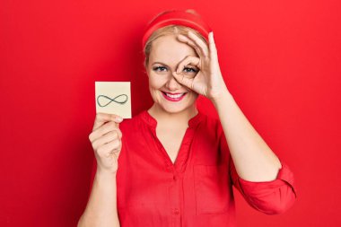 Sonsuz sembolü tutan genç sarışın kadın gülümseyen mutlu bir gülümseme. Tamam işareti yap. Elin parmaklarının arasından bak. 