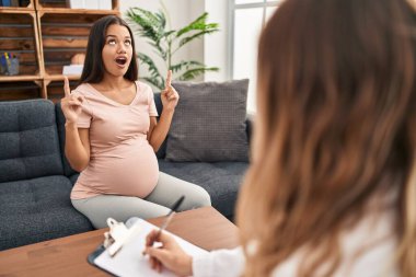 Young pregnant woman at therapy session amazed and surprised looking up and pointing with fingers and raised arms. 