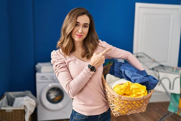 Young woman holding laundry basket pointing with hand finger to the side showing advertisement, serious and calm face