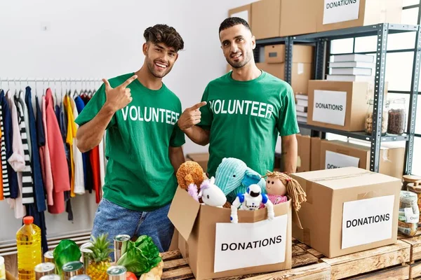 Young gay couple wearing volunteer t shirt at donations stand cheerful with a smile on face pointing with hand and finger up to the side with happy and natural expression
