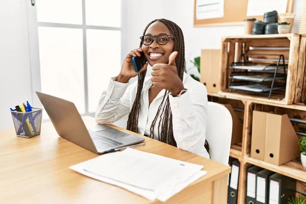 Black woman with braids working at the office speaking on the phone doing happy thumbs up gesture with hand. approving expression looking at the camera showing success.