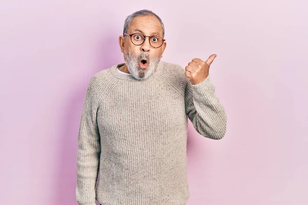 Handsome senior man with beard wearing casual sweater and glasses surprised pointing with hand finger to the side, open mouth amazed expression.