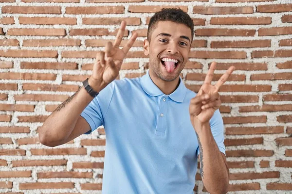 レンガの壁の上に立っているブラジルの若者は 両方の手の指を示す舌で笑みを浮かべて勝利の兆候を行う — ストック写真