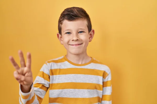 自信と幸せ笑みを浮かべている間 若い白人の子供は黄色の背景の上に立って指の数3を示すと指摘 — ストック写真