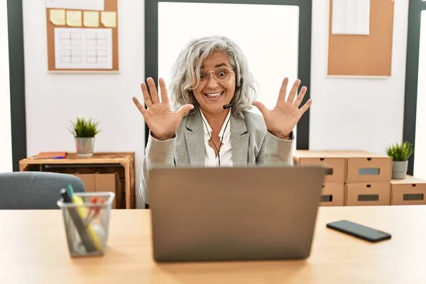 Middle age businesswoman sitting on desk working using laptop at office showing and pointing up with fingers number ten while smiling confident and happy.
