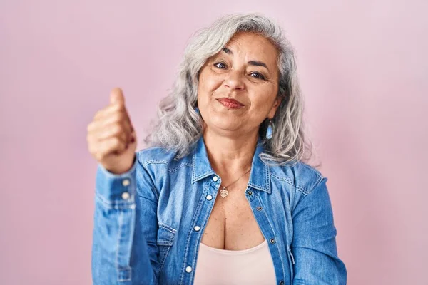 Middle age woman with grey hair standing over pink background looking proud, smiling doing thumbs up gesture to the side