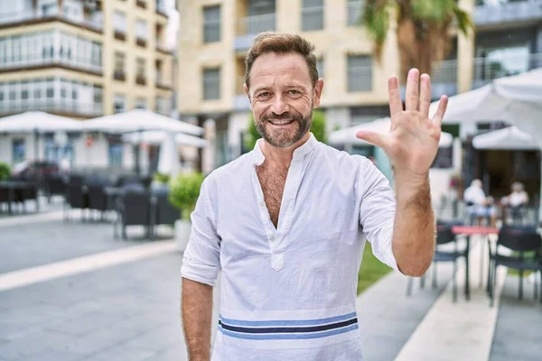 Middle age man outdoor at the city showing and pointing up with fingers number five while smiling confident and happy.