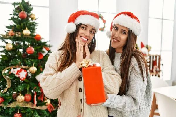 Two women surprise with gift standing by christmas tree at home