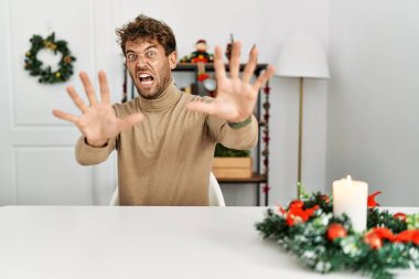 Masada sakallı genç ve yakışıklı bir adam oturuyor. Yılbaşı süslemeleri yüzünden korkmuş ve korkudan dehşete düşmüş. El hareketlerini bırak, şok içinde bağır. panik konsepti. 