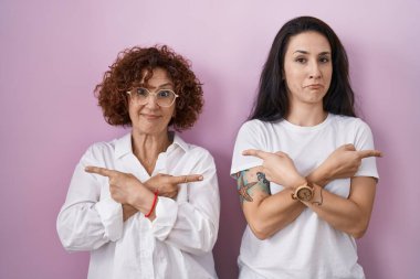İspanyol anne ve kızı pembe arka plan üzerine gündelik beyaz tişört giyip iki tarafı da parmaklarıyla işaret ediyorlar. Farklı yönler farklı. 