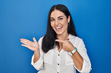 Mavi arka planda duran genç İspanyol kadın kameraya hayret ediyor ve gülümsüyor.. 