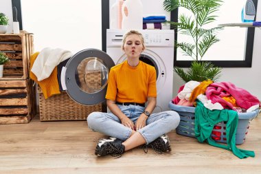 Young blonde woman doing laundry sitting by washing machine making fish face with lips, crazy and comical gesture. funny expression. 