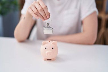 Young woman inserting house key on piggy bank at home