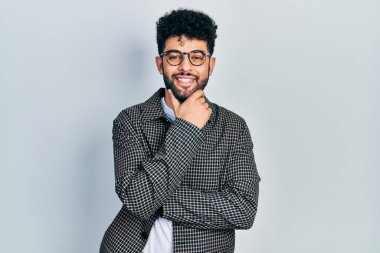 Sakallı, gözlüklü, kameraya kendinden emin, kolları çapraz ve çenesi açık gülümseyen genç bir Arap. Olumlu düşünmek. 