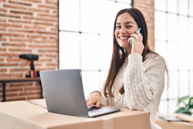 Genç bir kadın yeni evinde bilgisayarla akıllı telefondan konuşuyor.