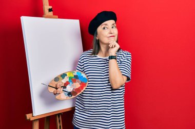 Middle age hispanic woman standing drawing with palette by painter easel stand with hand on chin thinking about question, pensive expression. smiling and thoughtful face. doubt concept. 