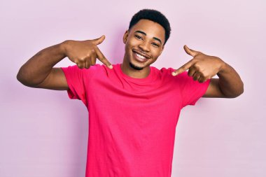 Genç Afrikalı Amerikalı adam Casua tişörtü giyiyor. Kendine güveni tam görünüyor. Kendini gururlu ve mutlu bir şekilde gösteriyor.. 