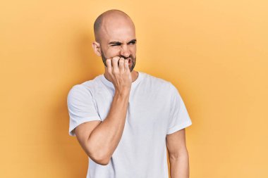 Gündelik beyaz tişört giyen genç kel adam gergin ve gergin görünüyor. Elleri ağızdan tırnak yiyor. anksiyete. 