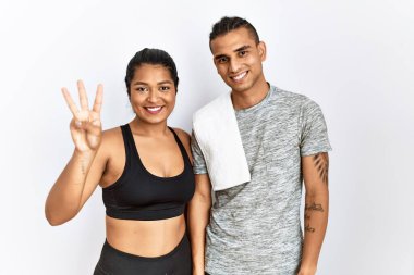 Spor kıyafetleri giyen genç bir çift izole edilmiş arka planda durup üç numaralı parmaklarıyla işaret ederken kendinden emin ve mutlu bir şekilde gülümsüyor.. 