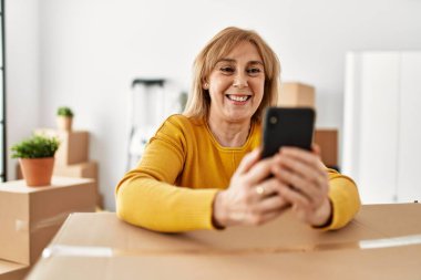 Orta yaşlı sarışın kadın yeni evinde akıllı telefon kullanarak mutlu bir şekilde gülümsüyor..